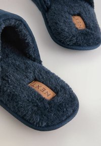 Navy blue slippers with fuzzy texture and soft lining, featuring a brown leather tag labeled "NEXT." Rounded shape and stitch detailing.