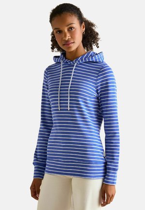 Young woman with curly hair wearing a blue and white striped hoodie and light-colored pants, standing against a plain background.