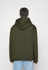 Felpa con cappuccio verde oliva dal taglio rilassato, con tasca a marsupio e polsini a costina, indossata con jeans azzurri chiari.
