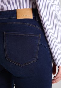 Mörkblå denimjeans med en bakficka, en tannfärgad läderlogotyp och kontrasterande gula sömmar. En randig skjorta är delvis synlig.