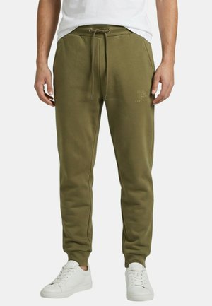 Homme portant un pantalon de survêtement vert olive avec cordon de serrage et des baskets blanches, debout devant un fond uni.