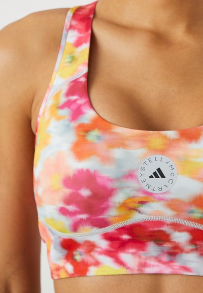 Spordirinnahoidja, millel on mitmevärviline lilleline muster roosa, oranži ja kollasega. Sisaldab Adidas'i logo silti ning sileda ja veniva kanga.