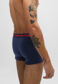 Marineblå boxershorts med rødt elastisk liv med "HUGO"-merke. Bløtt stoff og tettsittende design. Synlige tatoveringer på armen.