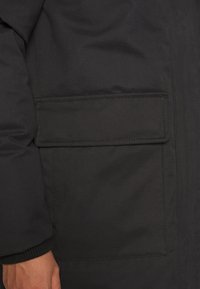 Gros plan sur la poche d'une veste noire avec un rabat couvrant l'ouverture et la main d'une personne partiellement visible près du poignet.