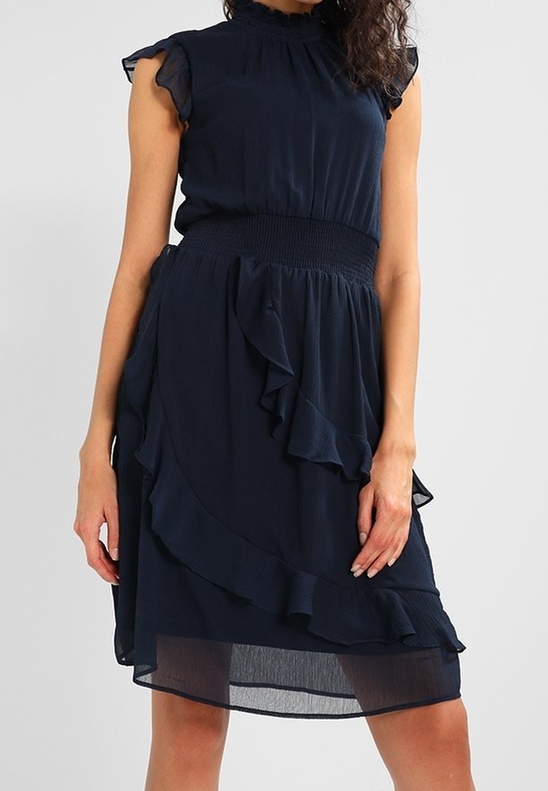 Femme portant une robe sans manches bleu marine avec des détails à volants et une taille froncée, debout devant un fond clair uni.