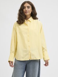 Gul knapp-skjorte laget av bomull med avslappet passform, med krage og lange ermer, sammen med lyseblå vide-jeans.