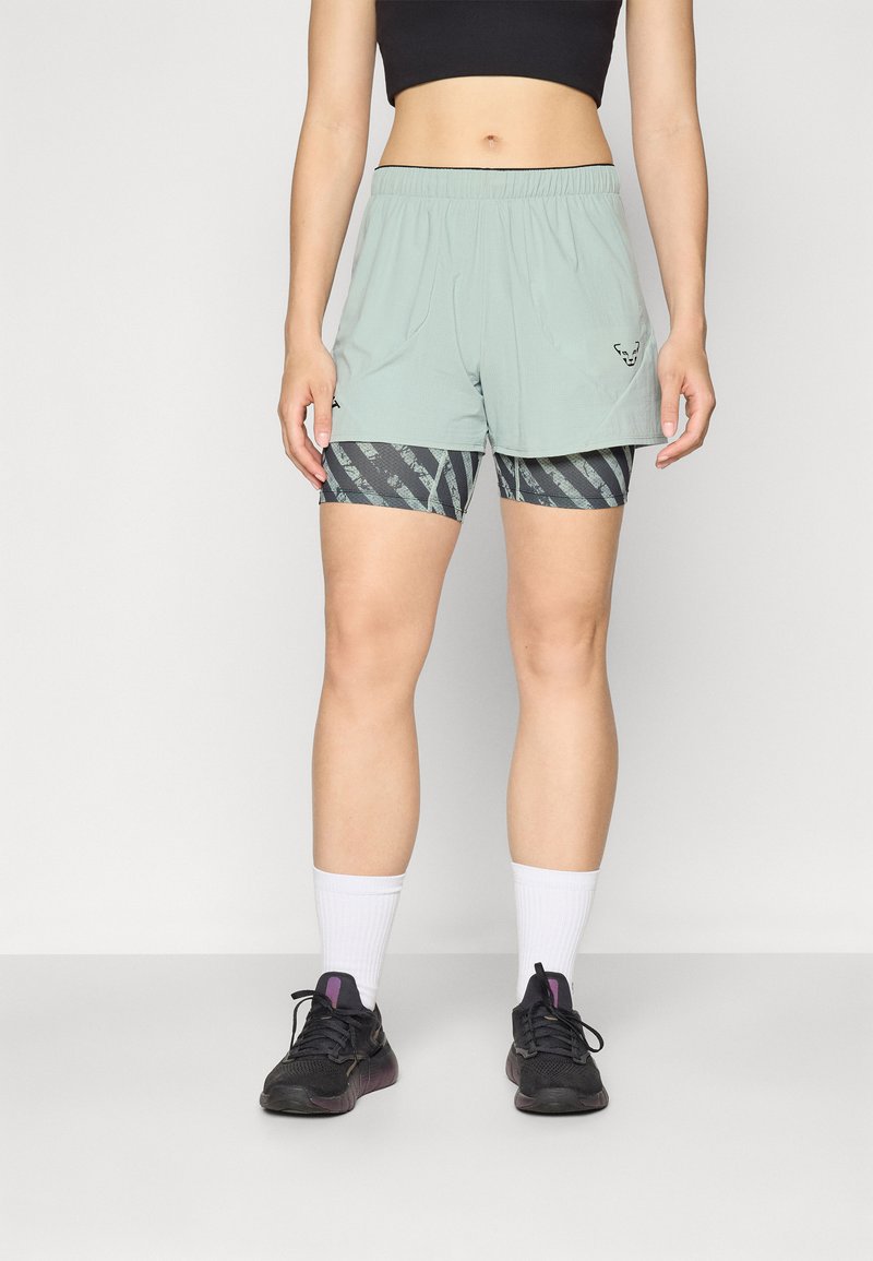 Lys grønne treningsshorts med elastisk midje, parret med svarte og grå mønstrede kompresjonsshorts under. Svarte treningssko.
