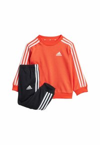 Felpa arancione con logo bianco Adidas e tre strisce bianche sulle spalle; abbinata a pantaloni neri con strisce simili.