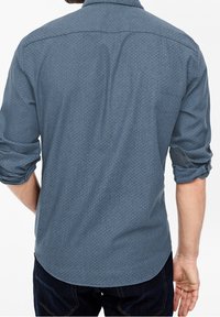 Hombre con las mangas remangadas que lleva una camisa de manga larga azul con estampado y jeans oscuros, visto desde atrás sobre un fondo blanco.