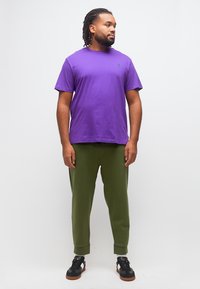 Άνδρας με πλεξουδάκια στα μαλλιά, που φοράει μοβ κοντομάνικο μπλούζα, ελιάς πράσινο παντελόνι και μαύρα αθλητικά παπούτσια, στέκεται και κοιτάζει προς το πλάι μπροστά σε ένα απλό φόντο.