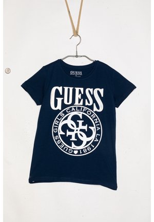 Mørkeblå T-shirt med korte ærmer med hvidt Guess-mærkelogo og cirkulær tekstdesign, hængende i et reb på en væg.