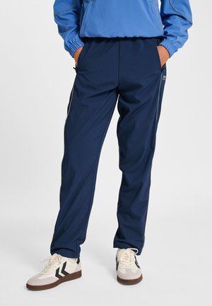 Personne portant un pantalon de survêtement bleu marine avec des fermetures éclair sur les côtés, assorti à des baskets blanches avec des rayures chevron noires, les mains dans les poches.