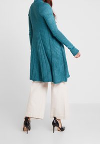 Teal lång kofta med vertikal ribbning, veckad rygg och åtsittande ärmar. Bärs över krämfärgade vida byxor och svarta högklackade skor.