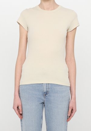 Mujer con una camiseta ajustada beige de manga corta y canalé, combinada con vaqueros de tiro alto azul claro, de pie frente a un fondo liso.