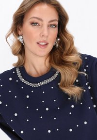 Tengerészkék pulóver gyöngydíszítéssel és kristályokkal díszített nyakkivágással. A modell feltűnő fülbevalót visel. Simább anyag és laza szabás.