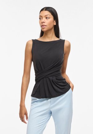 Vrouw draagt een mouwloze zwarte top met plooien en lichtblauwe broek, staand met één hand in de zak tegen een effen witte achtergrond.