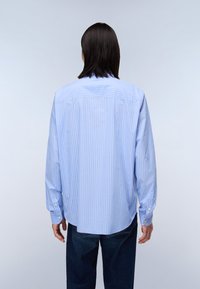Chemise rayée bleu clair et blanche, à manches longues, avec des poignets à boutons, un ourlet arrondi, fabriquée dans un tissu lisse. La vue de dos montre une coupe décontractée.