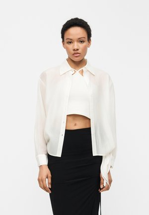 Junge Frau trägt ein transparentes weißes Hemd über einem weißen Croptop und einem hoch taillierten schwarzen Rock, steht vor einem schlichten Hintergrund.