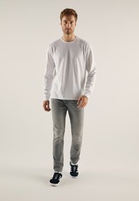 Chemise blanche à manches longues, jeans gris clair, baskets noires avec accents blancs. Le mannequin se tient sur un fond clair, mettant en valeur des vêtements décontractés.