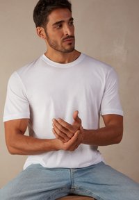 T-shirt en coton blanc avec un col rond et des manches courtes, associé à un jean en denim bleu. Modèle assis, les mains jointes.