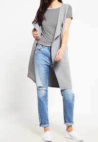 Cárdigan gris sin mangas sobre una camiseta de rayas blacas y negras, combinado con jeans claros desgastados y zapatillas blancas sin cordones.