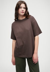 Hnědé bavlněné tričko s krátkým rukávem a volným střihem, s kulatým výstřihem; v centru má minimalistické logo.