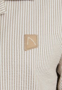 Camisa de tela a rayas en beige y crema, con un parche marrón y un logo blanco. La textura es suave y presenta finas líneas verticales.
