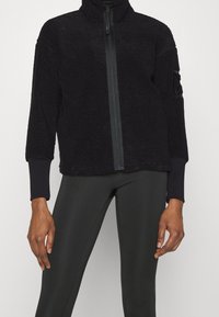 Czarna polarowa kurtka z wysokim kołnierzem, zamkiem błyskawicznym na całej długości z przodu oraz ściągaczami przy rękawach. Posiada teksturowaną powierzchnię i naszywkę na lewym rękawie.