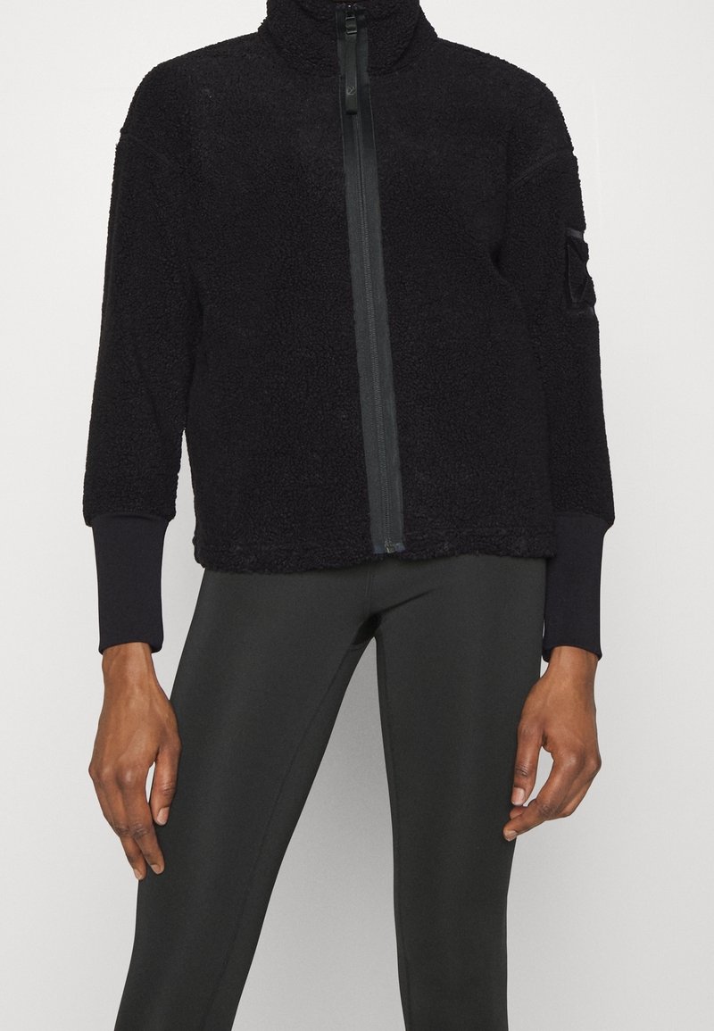 Czarna polarowa kurtka z wysokim kołnierzem, zamkiem błyskawicznym na całej długości z przodu oraz ściągaczami przy rękawach. Posiada teksturowaną powierzchnię i naszywkę na lewym rękawie.