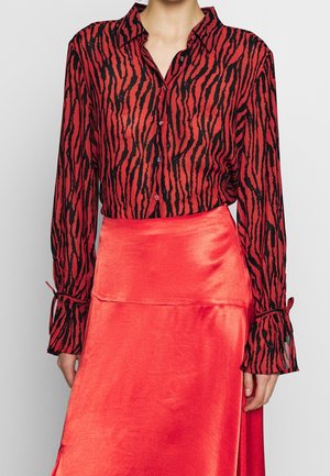 Kobieta w czerwono-czarnej bluzce w zwierzęcy wzór zapinanej na guziki z długimi rękawami oraz w jasnoczerwonej satynowej spódnicy.