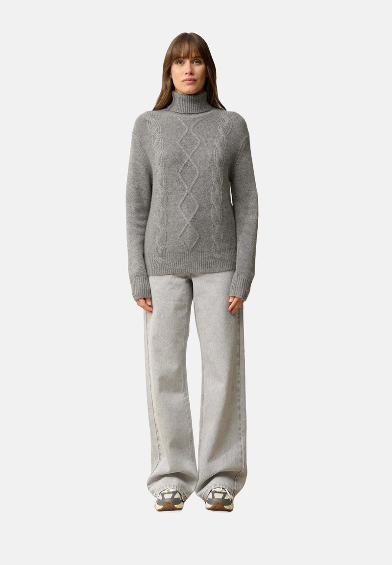 Szary sweter z dzianiny o wysokim dekolcie i wzorze w warkocze, w połączeniu z jasnoszarymi spodniami o szerokich nogawkach i wielokolorowymi sneakersami.