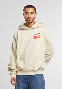 Kremfarget hettegenser med frontlomme, ribbede mansjetter og et logo-patch som viser "UpscaleSt" i kontrastfargene svart og rødt.