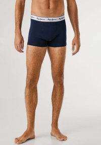 Mann trägt marineblaue Boxershorts mit weißem Bund der Marke "Pepe Jeans" und steht barfuß vor einem einfarbigen, hellen Hintergrund.