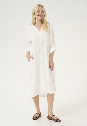 White knee-length dress with a buttoned placket, roll-up sleeves, and side pockets, made from a lightweight, textured fabric. Animal print flats.