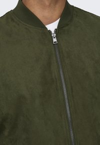 Giubbotto bomber verde oliva realizzato in morbido camoscio con chiusura frontale a zip, collo e polsini a coste e una texture liscia.
