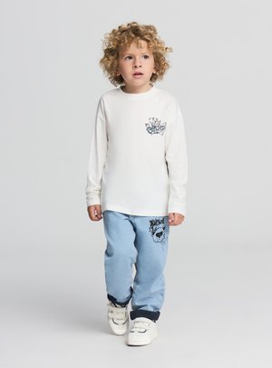 Kremowa koszula z długim rękawem z czarną grafiką na piersi. Jasnoniebieskie spodnie jeansowe z czarnym wzorem na jednej nogawce. Białe tenisówki na rzepy.