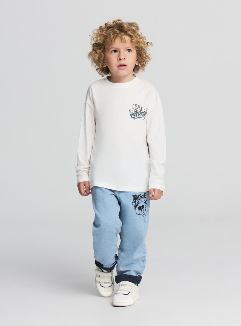 Kremowa koszula z długim rękawem z czarną grafiką na piersi. Jasnoniebieskie spodnie jeansowe z czarnym wzorem na jednej nogawce. Białe tenisówki na rzepy.