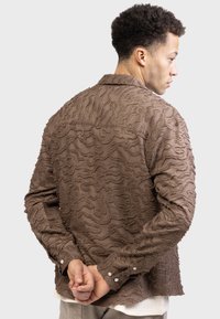 Brown textured shirt with a wave-like pattern, collar, long sleeves, and buttoned cuffs, featuring a relaxed fit and visible stitching details.