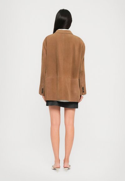 Donna con lunghi capelli scuri indossa un blazer marrone oversize, una mini gonna nera e mule bianche con tacco, in piedi di spalle contro uno sfondo neutro.