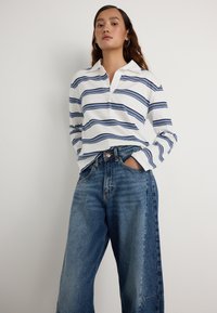 Bijela polo majica dugih rukava s tamnoplavim horizontalnim prugama, u kombinaciji s hlačama s visokim strukom i širokim nogavicama u plavoj boji, koje imaju kopčanje na dugme i džepove.