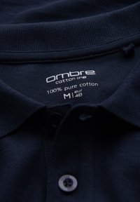 Mörkblå bomullspolo med krage och etikett som visar märket "ombre", 100% ren bomull, storlek M (EUR 48), och delvis synlig knapp.