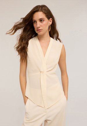 Top beige sans manches avec un col en V et un détail de nœud à l'avant. Présente une texture lisse et un ourlet fluide et asymétrique.