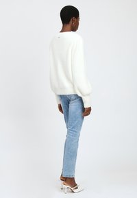 Een vrouw met kort haar staat met de rug naar ons toe en draagt een witte fluffy trui, lichtblauwe jeans en witte hoge hakken met riempjes.