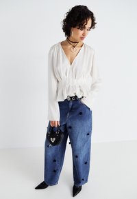 Bela peplum bluza z dolgimi rokavi, v kombinaciji z modrimi širokimi hlačami z visokim pasom, ki imajo črne cvetlične okraske. Drži črno torbico v obliki srca.