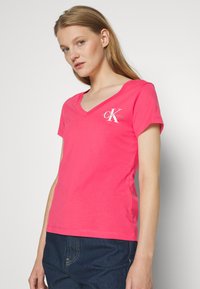 Camiseta de algodón rosa con cuello en V, mangas cortas y un logo blanco en el lado izquierdo del pecho. Textura suave y diseño ajustado.