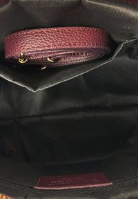 Wnętrze torby wyłożone czarną tkaniną z rulonowym paskiem z bordowej skóry o fakturze oraz dopasowaną metką przymocowaną wewnątrz.