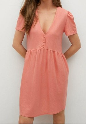 Frau trägt ein korallenrotes Kleid mit kurzen Ärmeln, tiefem V-Ausschnitt und drei Knöpfen am Oberteil, steht vor einem einfarbigen Hintergrund.