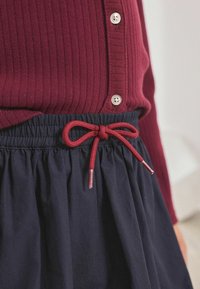 Bordeaux geribbelde trui met zilveren knopen, gecombineerd met een donkerblauwe rok met een gathered tailleband en een rode trekkoord strik met zilveren uiteinden.