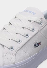 Άσπρο αθλητικό παπούτσι Lacoste με υφασμάτινο επάνω μέρος, λευκά κορδόνια και κεντημένο λογότυπο κροκόδειλου στο πλάι κοντά στη χοντρή λαστιχένια σόλα.
