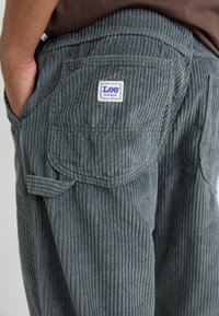 Gray corduroy pants with a ribbed texture, featuring a side pocket and a blue and white Lee Workwear label on the back.